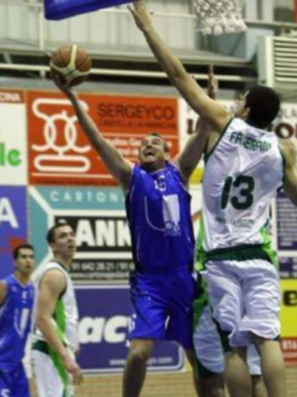 Jugadores de baloncesto en acción durante un partido, uno intenta marcar mientras el otro bloquea el balón.