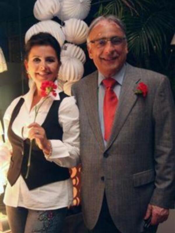 Dos personas posando juntas, una con un vestido blanco y negro y una corona de flores, la otra con un traje gris claro y un lazo rojo, frente a una decoración de globos blancos y plantas verdes.