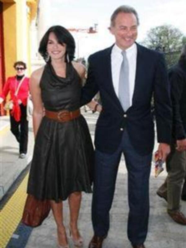 Una pareja formal caminando juntos en un entorno urbano, con el hombre vestido con traje y corbata y la mujer con vestido negro y cinturón.