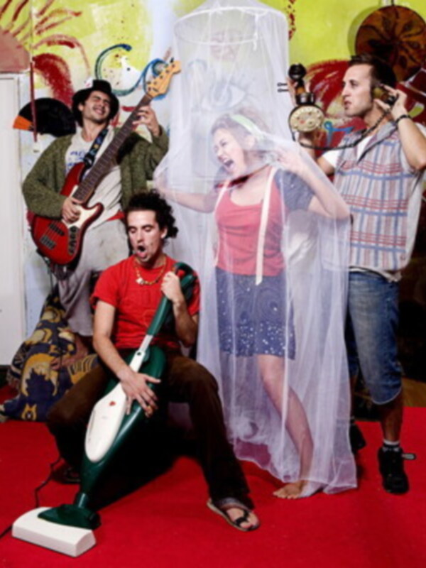 Un grupo de músicos en un escenario con instrumentos y una mujer vestida con una malla de seda en el centro, rodeados de un ambiente vibrante y colorido.