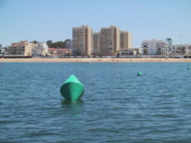 Bote de color verde flotando en el agua, con edificios y árboles en el fondo.