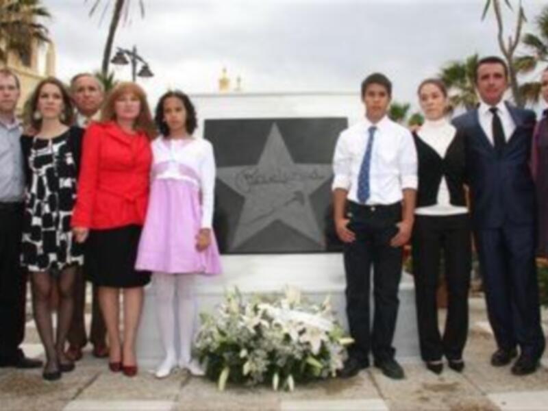 Una foto de grupo de personas en una ceremonia formal, con una estrella en el centro y una ofrenda floral en el frente. La escena parece ser en un lugar público con vegetación en el fondo.
