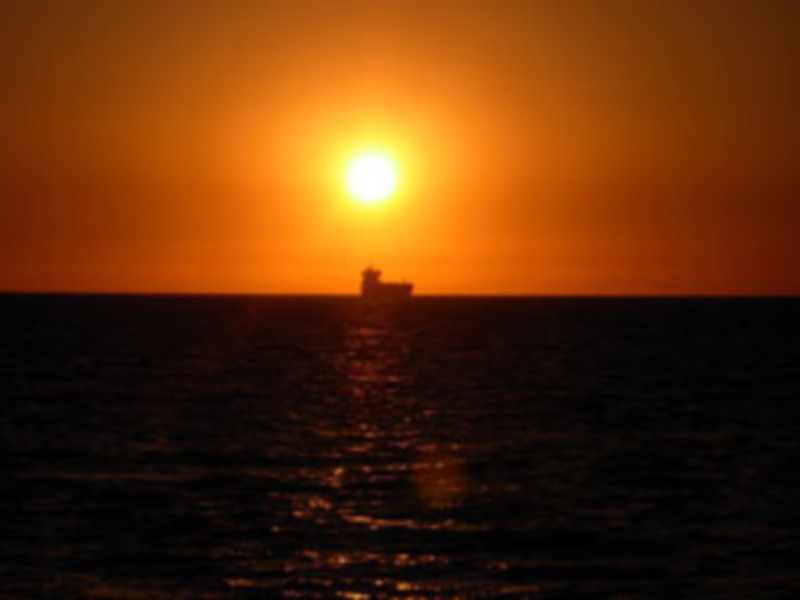 Puesta de sol sobre el océano, con un barco en el horizonte. El sol se oculta detrás de la montaña, iluminando el cielo de naranja y rojo. La imagen captura la calma y la belleza del momento.