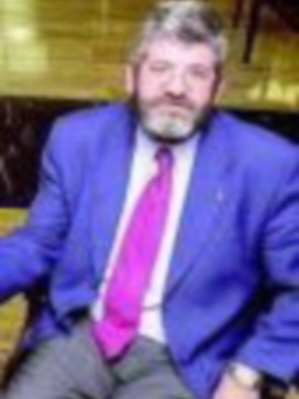 Hombre en traje con corbata rosa, sentado en una silla de madera, fondo de madera y cristal.