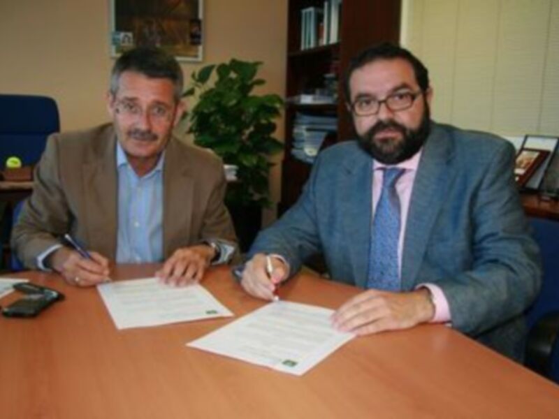 Dos hombres sentados en una mesa, uno con una chaqueta de lana y el otro con una camisa azul, firmando documentos en un entorno de oficina con plantas y libros en el fondo.