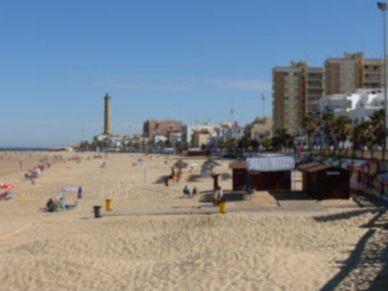 Paisaje costero con playa, edificios y faro en el horizonte bajo un cielo azul claro.
