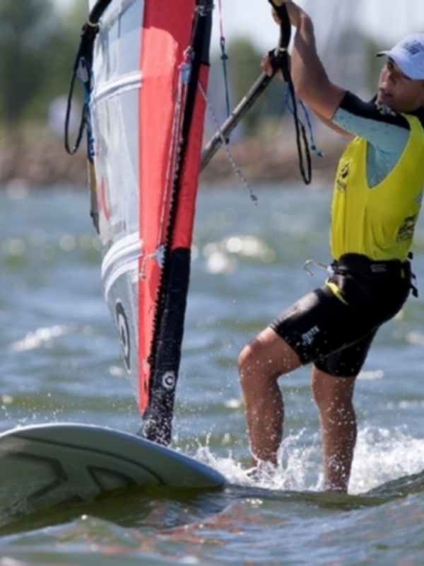 Un windsurfer en acción, con una vela roja y negra y un chaleco amarillo, en un mar tranquilo con pequeñas olas.