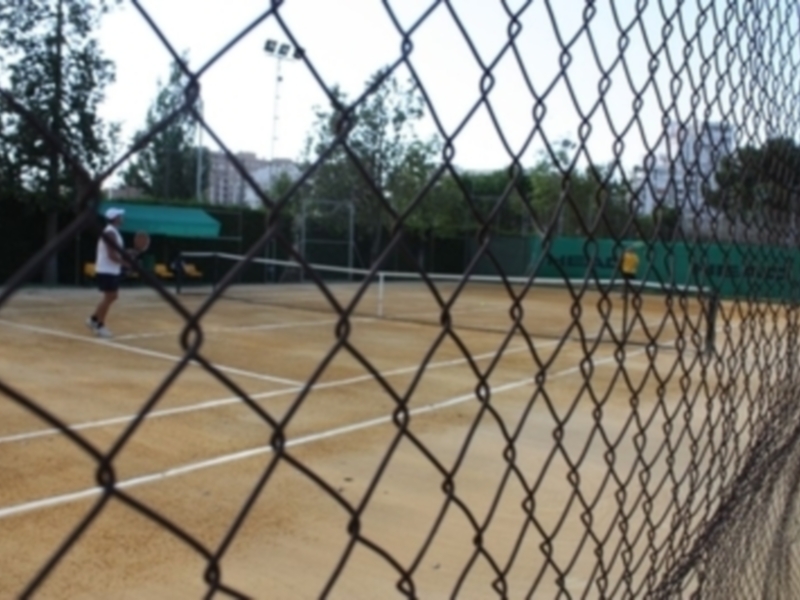 Jugador de tenis en acción en una cancha de tierra batida, vista a través de una cerca de alambre.
