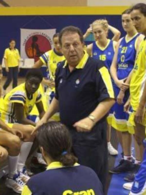 Equipo femenino de baloncesto en una cancha de competición, con un entrenador en el centro y jugadoras de diferentes equipos en el fondo.