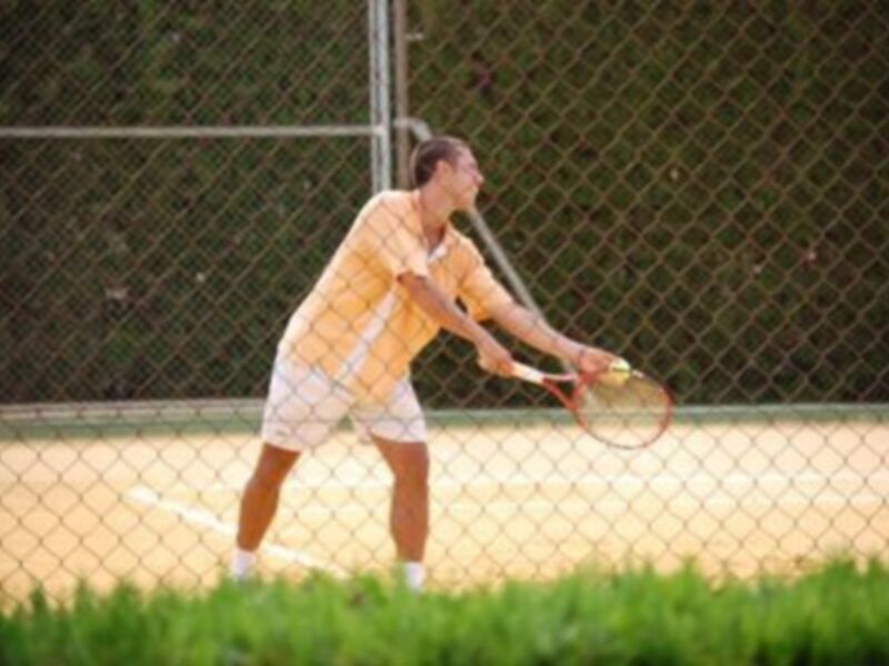 Un hombre jugando tenis en un campo de tenis con una cancha de color amarillo y una cerca de metal en el fondo.