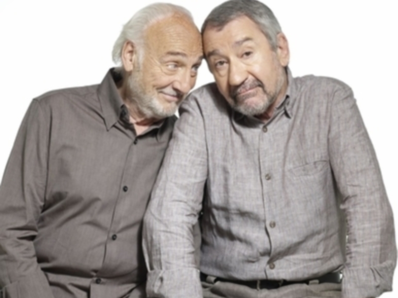 Dos hombres mayores sonrientes, uno con barba y el otro con bigote, posando juntos en una imagen de fondo blanco.