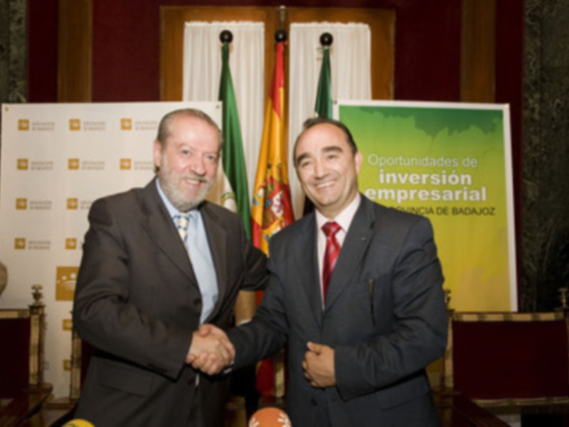 Dos hombres en traje se saludan en un evento de inversión empresarial, ante un fondo con banderas y letreros de España y Andalucía.