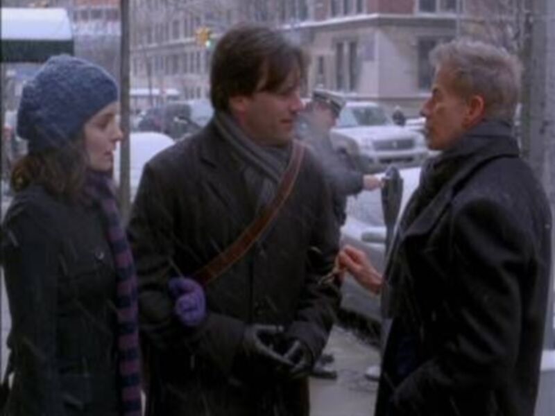 Tres personas conversan en una calle nevada, vestidas con abrigos y bufandas, en un ambiente urbano.