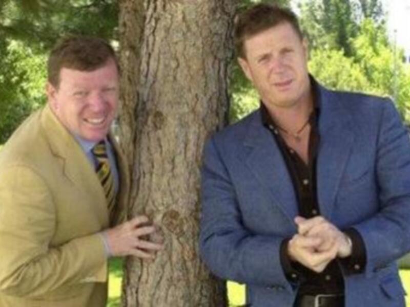 Dos hombres sonrientes posando junto a un árbol, uno de ellos con una camisa blanca y el otro con una chaqueta azul.