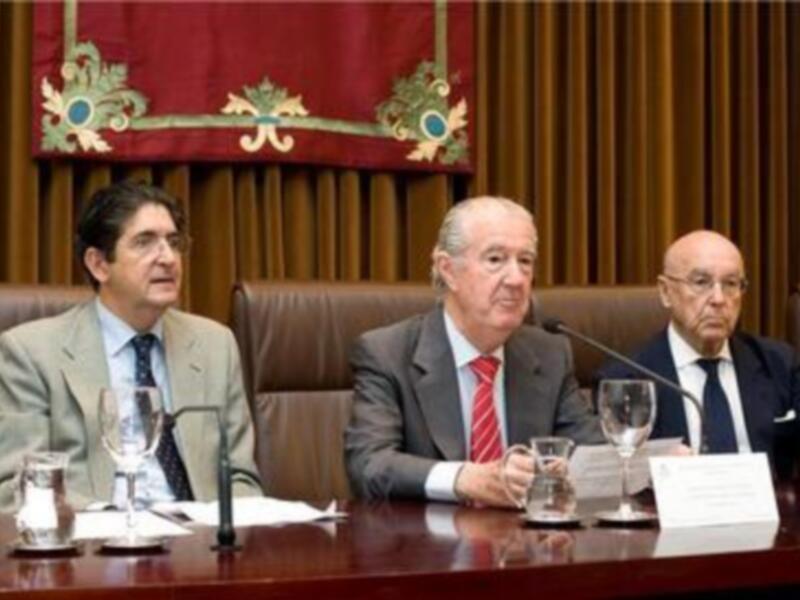 Tres hombres sentados en una sala de conferencias, con un fondo de cortinas y un escudo ceremonial en el marco.