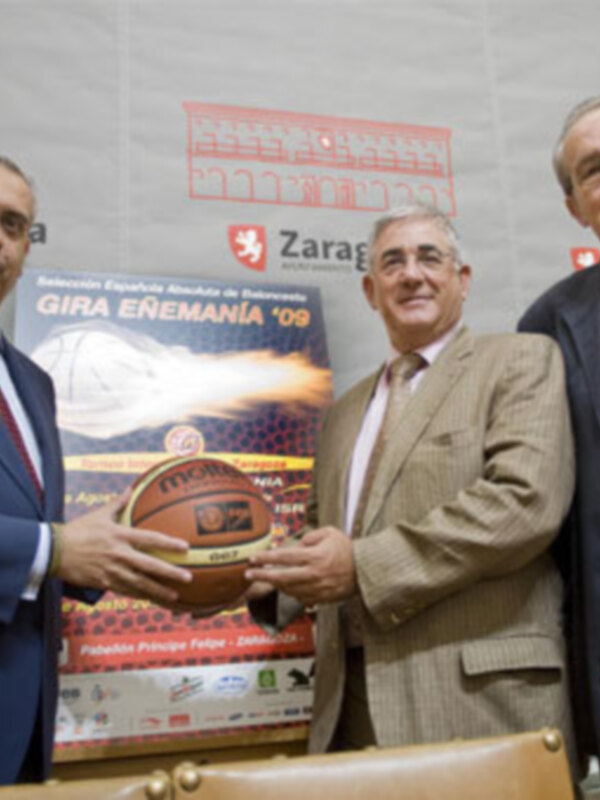 Tres hombres en una reunión, uno de ellos sostiene un balón de baloncesto. En el fondo, hay una pancarta con el nombre 'Zaragoza' y un logo con la palabra 'Gipuzkoa'.
