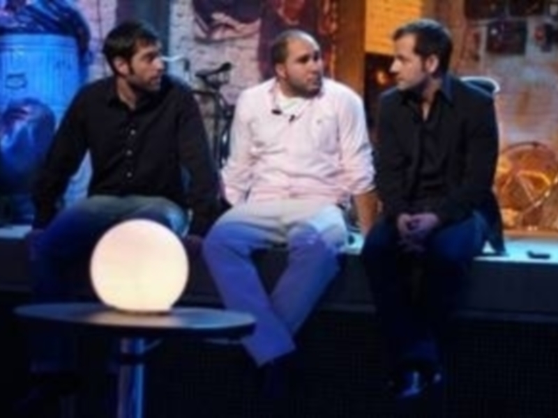Tres personas sentadas en un set de televisión, con un fondo que parece ser una pantalla de televisión con imágenes borrosas.