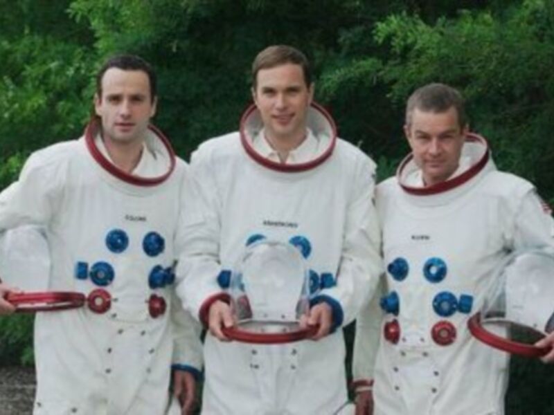 Tres astronautas en trajes espaciales sosteniendo consolas de control, posando al aire libre.