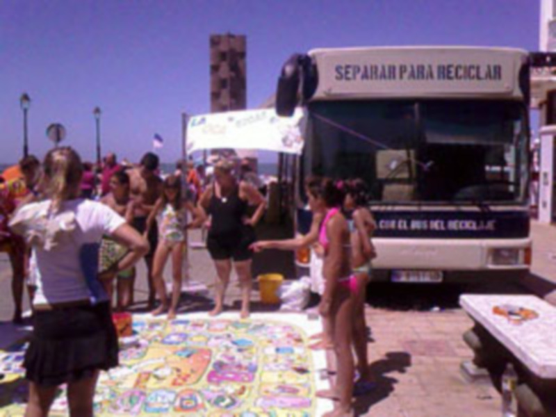 Fiesta de arte en la playa con gente pintando y un autobús de reciclaje en el fondo.