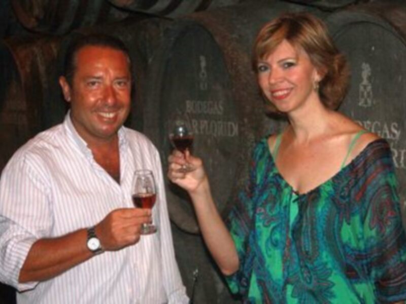 Dos personas disfrutan de un vino tinto en una bodega, rodeadas de barricas de roble.