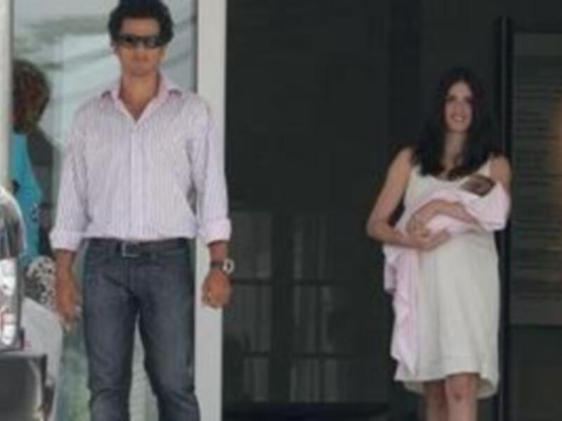Un hombre y una mujer caminan juntos, el hombre lleva una camisa de rayas y pantalones de mezclilla, mientras la mujer lleva una blusa blanca y un vestido largo. Ambos parecen estar en un evento formal.