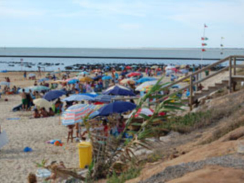 Paisaje de playa con gente disfrutando bajo sombrillas y en la arena, con el mar en el fondo y un faro en la distancia.