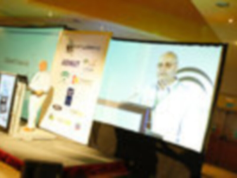 Una escena de conferencia con dos presentadores en una pantalla grande, sosteniendo un libro y un teléfono, respectivamente. El escenario está iluminado con luces brillantes y el fondo muestra una pantalla de proyección con imágenes de tecnología.