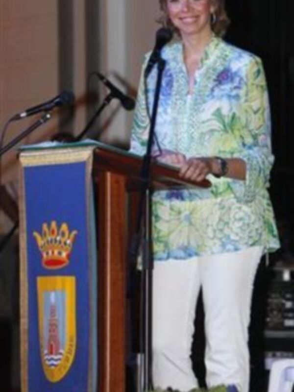 Una mujer en un traje floral y pantalones blancos está hablando en un podio con un escudo de armas en el fondo. El escudo tiene una corona y un león, lo que sugiere un contexto oficial o institucional.