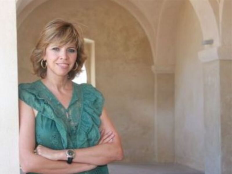Mujer con camisa verde y brazos cruzados en un entorno arquitectónico con arcos y paredes de color claro.