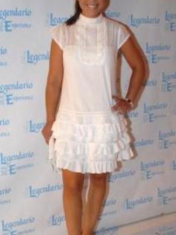 Una mujer con vestido blanco y sandalias, posando frente a un fondo con el logotipo 'Legendario' y 'E!'.