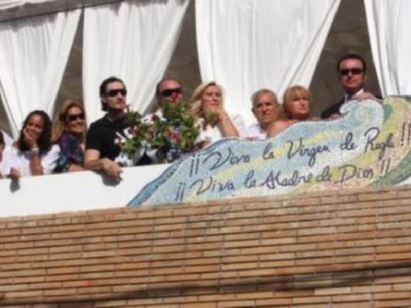 Una multitud de personas se reúne en un balcón, con una pancarta que dice "¡Viva la Virgen de Puga! ¡Viva la Madre de Dios!" en el centro. La imagen parece ser parte de una celebración religiosa o festiva.