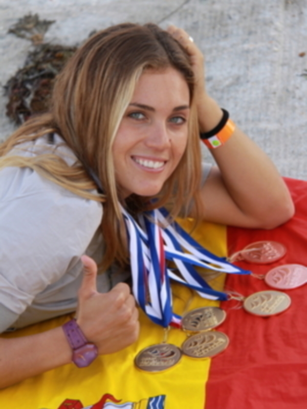 Una mujer sonríe mientras sostiene varias medallas de oro y plata, con una bandera española al fondo.