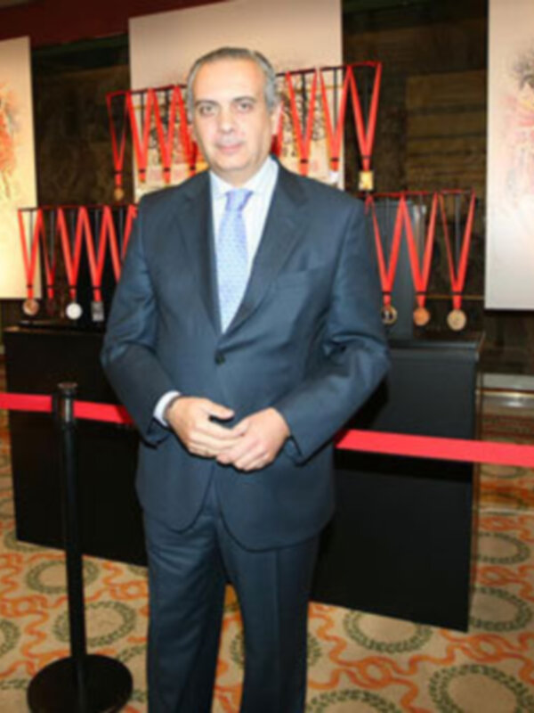 Hombre en traje negro y corbata azul posa en un evento formal con medallas y carteles en el fondo.