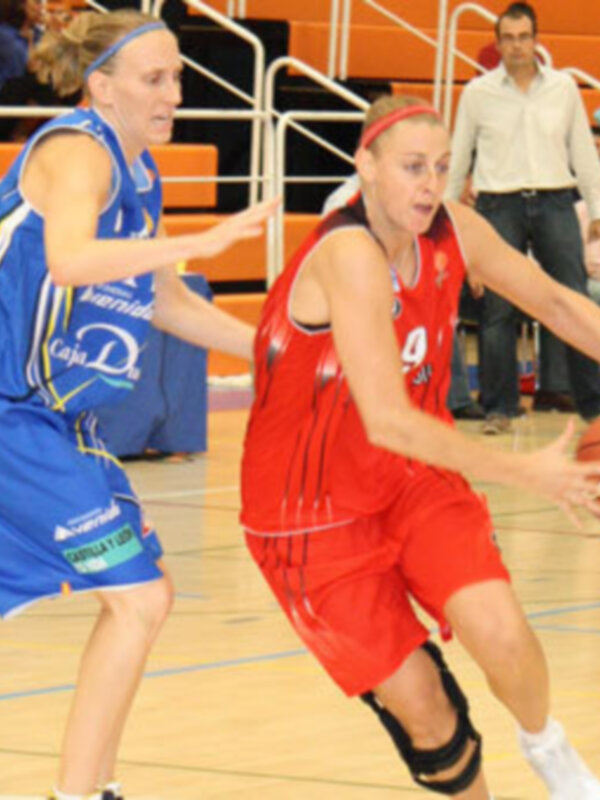 Dos jugadoras de baloncesto en acción durante un partido. La jugadora en azul lleva una camiseta con el logo 'Caja D' y la jugadora en rojo lleva una camiseta con el número 4. Ambas están en una cancha de baloncesto con una portería de hierro en el fondo.