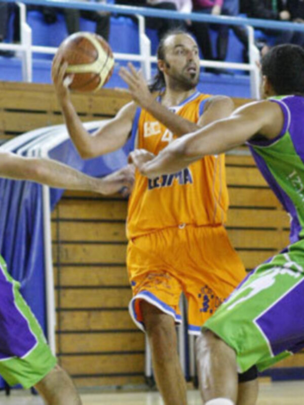 Jugadores de baloncesto en acción durante un partido, con un atleta en camiseta naranja driblando el balón mientras es marcado por jugadores en camisetas púrpuras y verdes.