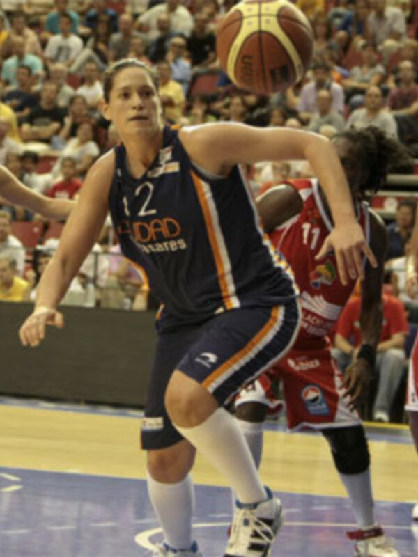 Jugadora de baloncesto en acción durante un partido, driblando el balón mientras es marcada por la defensa en un estadio lleno de espectadores.