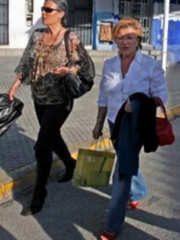 Dos mujeres caminan por una calle, una con bolso y la otra con bolsa de compras. La escena parece ser en un entorno urbano, posiblemente una ciudad o pueblo.