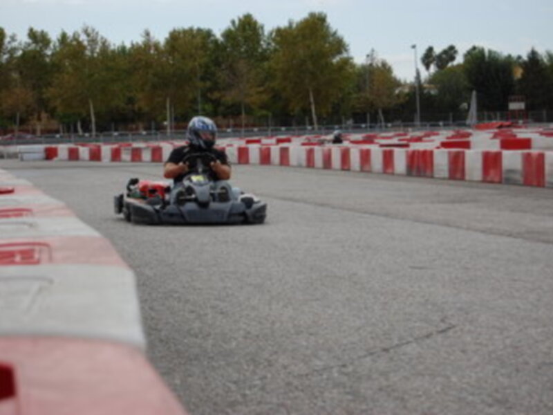 Un piloto en un kart de carreras en una pista de karting, con un fondo de árboles y un cielo despejado.