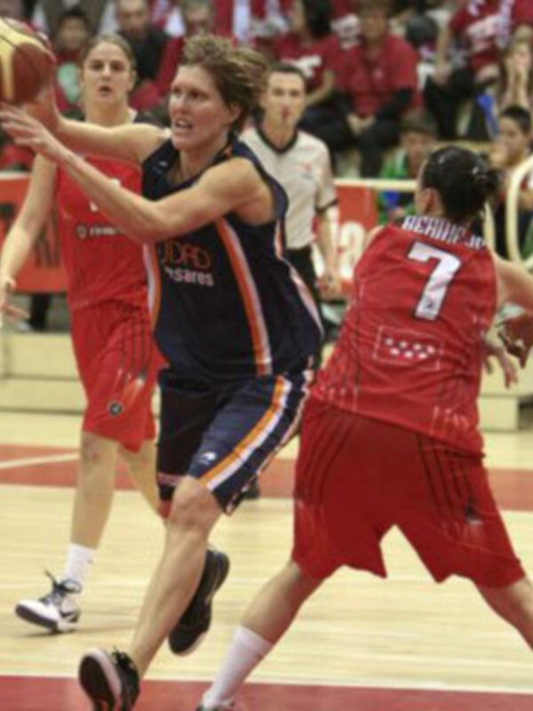 Jugadores de baloncesto en acción durante un partido, una jugadora en azul intenta lanzar mientras es marcada por una jugadora en rojo.