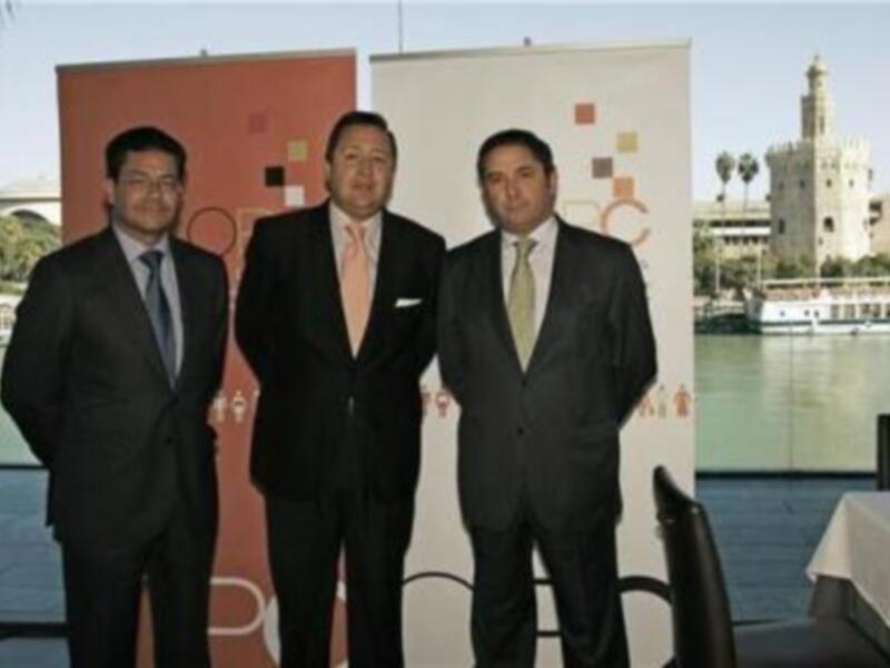 Tres hombres en trajes posan juntos en un evento profesional frente a un cartel y un paisaje urbano con una torre histórica al fondo.