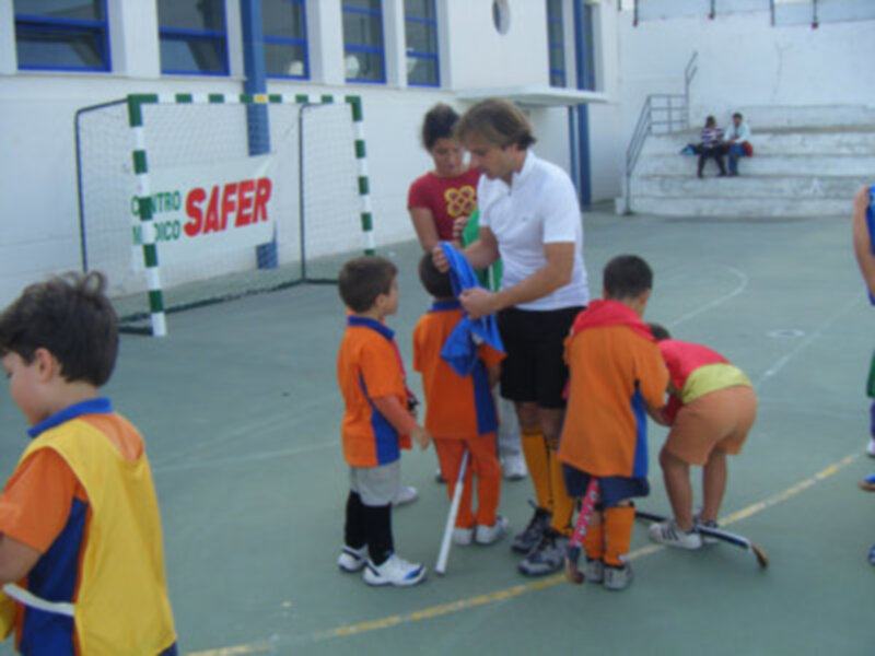 Niños jugando hockey en un campo de fútbol, con una portería de seguridad visible en el fondo.