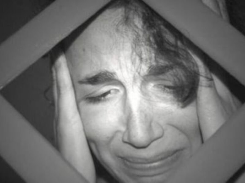 Una persona con una expresión de dolor o preocupación, cubriendo su rostro con las manos, detrás de una estructura metálica.