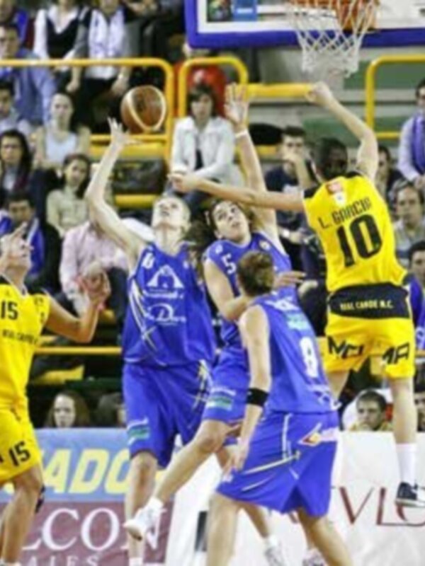Jugadores de baloncesto en acción durante un partido, con el aro y la canasta visible en el fondo.