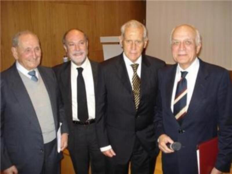 Cuatro hombres en trajes posan juntos en una sala de conferencias, uno de ellos sostiene un micrófono.