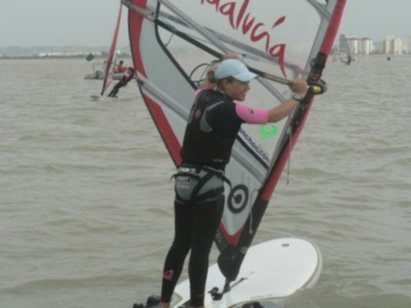 Una persona practicando windsurf en el agua, con una vela roja y blanca en el fondo.