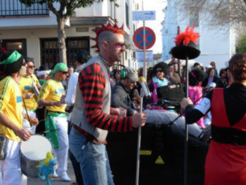Una persona con un sombrero de cuerno rojo y una camiseta a rayas rojas y negras, posando en una calle con otros personajes y un árbol en el fondo.