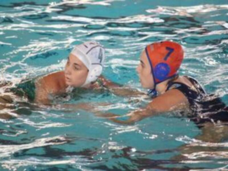 Dos jugadores de waterpolo en acción en el agua, uno con casco blanco y otro con casco rojo y azul.