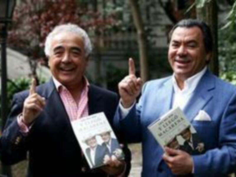 Dos hombres sonrientes sostienen libros y ponen el dedo en el aire en un entorno al aire libre.