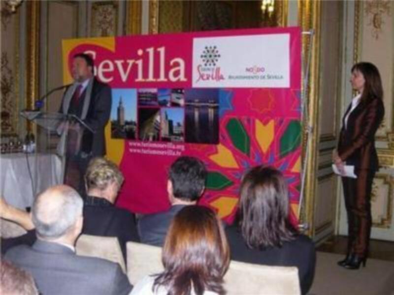 Presentación en Sevilla: hombre hablando al micrófono con audiencia y cartel de la ciudad en el fondo.