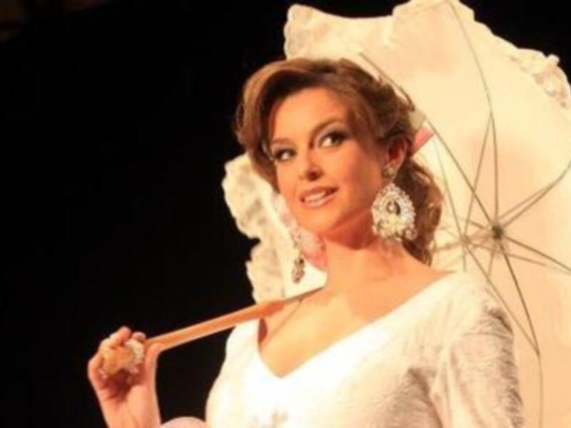 Una mujer en un vestido blanco sostiene un bastón de madera, posando con elegancia en un entorno festivo.